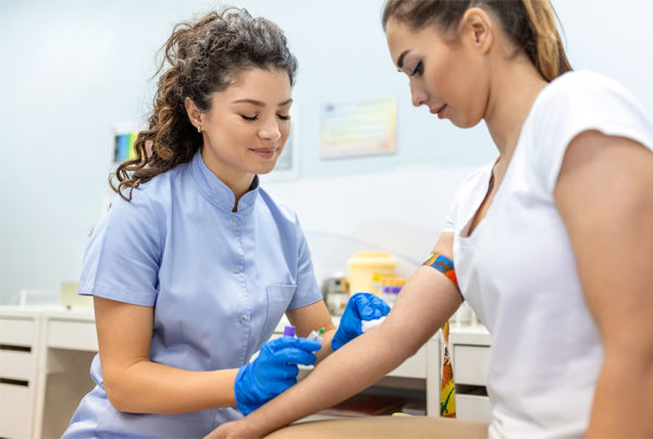 An image depicting a female phlebotomist wearing scrubs and gloves preparing to draw blood from another female who is the patient
