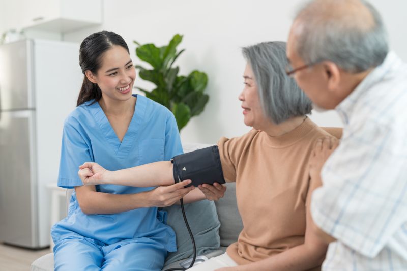 Asian caregiver doctor using digital pressure gauge measuring blood pressure of elderly woman patient Young female nurse in uniform gives consultation during home visit.