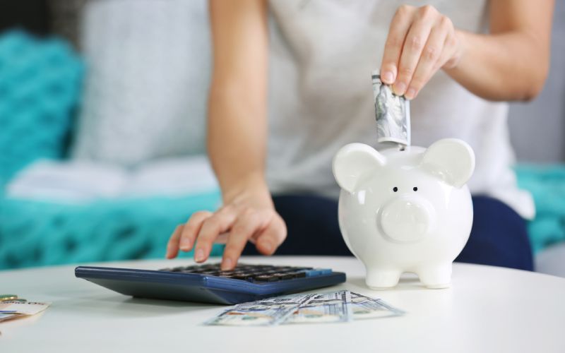 Female hand putting money into piggy bank and counting on calculator closeup