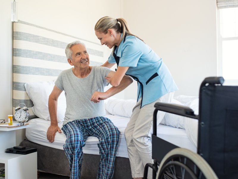 Smiling nurse assisting senior man to get up from bed. Caring nurse supporting patient while getting up from bed and move towards wheelchair at home. Helping elderly disabled man standing up.