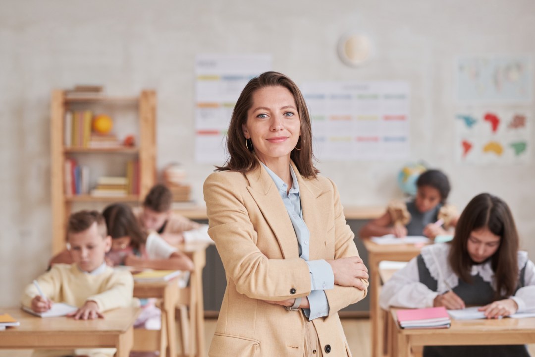 Waist up portrait of smiling female teacher looking at camera while posing confidently standing with arms crossed in school classroom, copy space